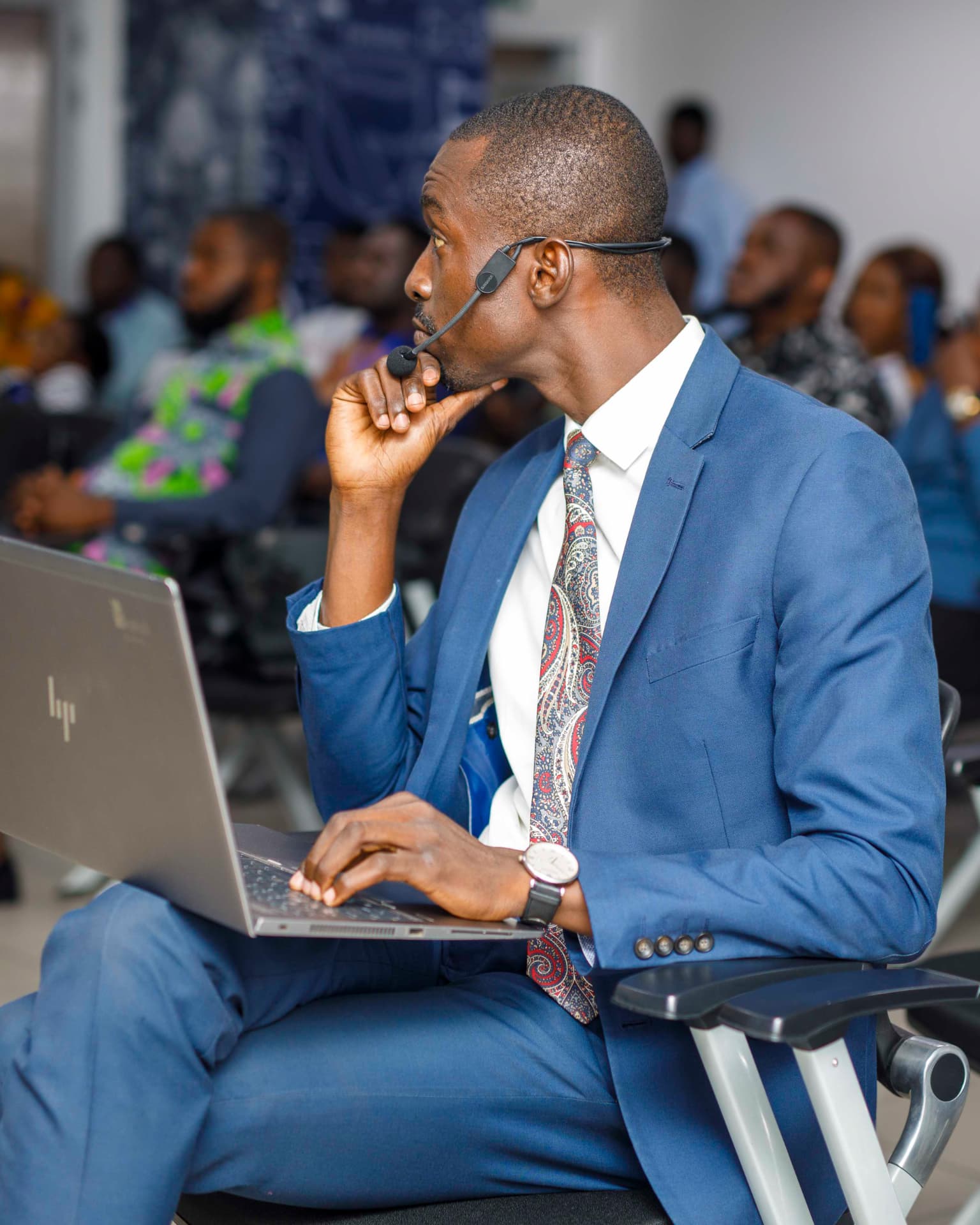 King David Amo seated in a blue suit with a headset microphone and laptop at a professional event